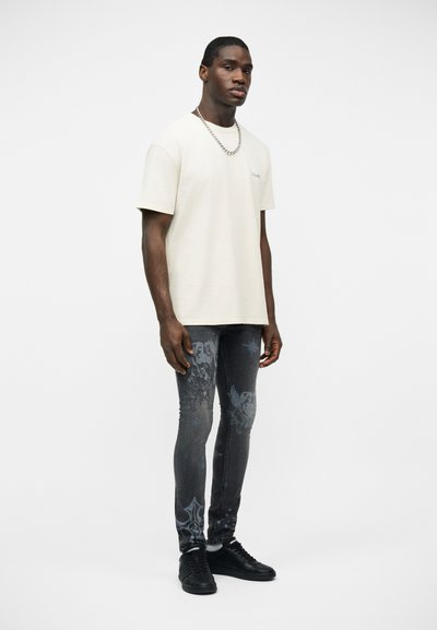 Άνδρας που φοράει λευκό κοντομάνικο μπλουζάκι, σκουρόχρωμο στενό τζιν με σχέδια, μαύρα αθλητικά παπούτσια και ασημένιο κολιέ αλυσίδα, στέκεται μπροστά σε λευκό φόντο.