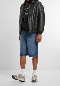 Černá kožená bomber bunda, černá mikina s potiskem, modré denimové šortky, bílé ponožky, šedé a béžové atletické boty. Neformální streetwear vzhled.