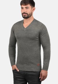 Grå V-hals sweater lavet af blødt stof, med ribstrikket kant og ærmer, samt en lille læderetiket på nederste venstre side.