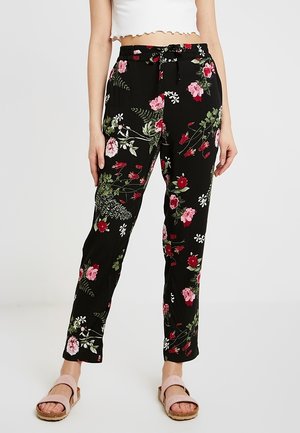Schwarze Hose mit pinkem, rotem und weißem Blumenmuster, gebundene Taille, kombiniert mit einem weißen bauchfreien Top und pinken Sandalen mit offenem Zehenbereich.