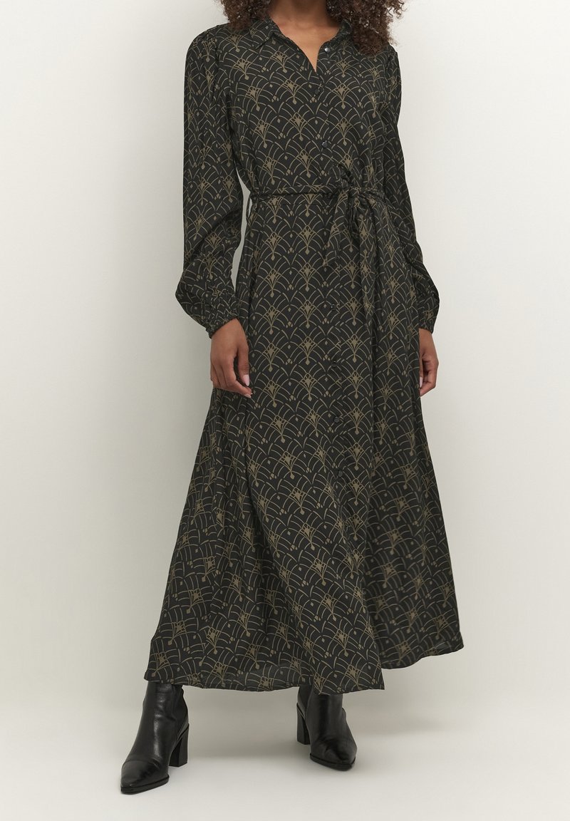Femme portant une robe longue noire avec des motifs géométriques dorés, manches longues, taille nouée et bottines noires à talons.