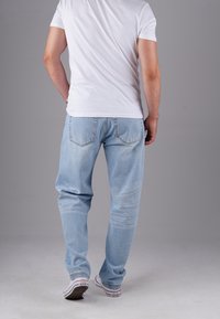 Hombre con camiseta blanca, jeans de un azul claro y zapatillas Converse blancas de pie de espaldas a la cámara sobre un fondo gris.