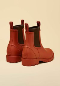 Stivali Chelsea rossi in gomma con superficie texturizzata, pannelli elastici laterali marroni e linguette per indossarli. Suole spesse e sagomate per una migliore aderenza.