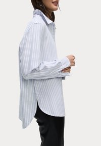 Camisa abotonada azul y blanca a rayas con cuello, mangas largas, dobladillo curvado y detalles de botones en los lados. Textura de algodón suave.