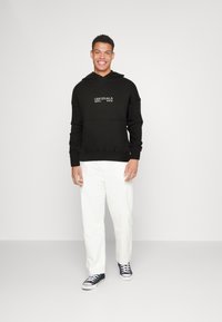Felpa nera con cappuccio e tasca frontale, logo "ORIGINALS" in bianco. Abbinata a pantaloni bianchi e sneakers nere. Look e design casual.