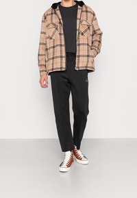 Person som bär en beige och orange rutig zip-jacka, svart t-shirt, svarta dragsko-byxor och högskor med bruna, vita och marinblå paneler.