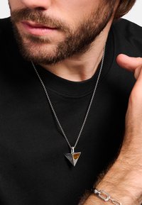 Silberanhänger in dreieckiger Form mit Bernsteinakzent, an einer Kette getragen, über einem schwarzen Hemd getragen, mit strukturierten und glatten Oberflächen.