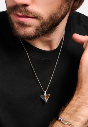 Silberanhänger in dreieckiger Form mit Bernsteinakzent, an einer Kette getragen, über einem schwarzen Hemd getragen, mit strukturierten und glatten Oberflächen.