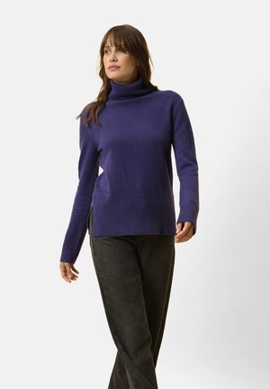 Purple turtleneck sweater in soft, textured fabric. Features long sleeves, side slits, and a relaxed fit, paired with dark jeans.