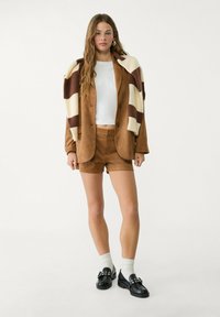 Braune Wildleder-Blazer und Shorts mit einem cremefarbenen und braun gestreiften Strickschal, kombiniert mit schwarzen Slippern und weißen Socken.
