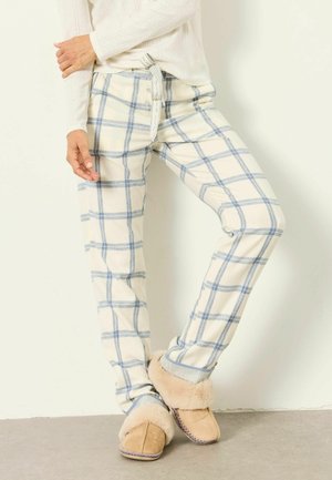 Persona che indossa pantaloni a quadri crema e blu con cintura in vita e pantofole beige foderate in pelliccia, in piedi contro un muro bianco.