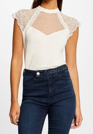 Vrouw draagt een crèmekleurige kanten top met kapmouwen en een doorzichtige stippenbovenzijde, en donkere high-waisted jeans. Romp zichtbaar, armen ontspannen langs het lichaam.