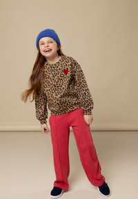 Glunderend meisje met een blauwe gebreide muts, een trui met luipaardprint en een rode hart, rode broek en marineblauwe sneakers, staand tegen een beige achtergrond.