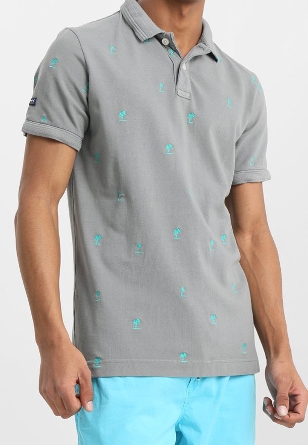 Homme portant un polo gris avec de petits motifs de palmiers turquoise et un short bleu vif sur un fond uni.
