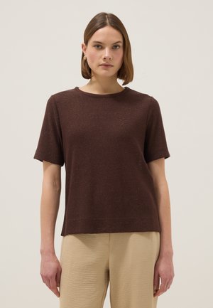 Vrouw met kort bruin haar, gekleed in een bruin shirt met korte mouwen en beige broek, staand tegen een effen lichte achtergrond.