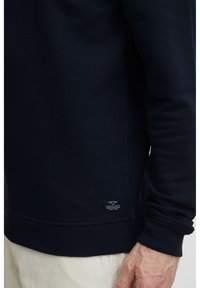 Felpa navy con polsini e orlo a costine. Presenta un'etichetta cucita sull'orlo inferiore. Texture liscia, vestibilità casual e maniche lunghe.