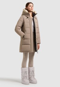 Béžová péřová bunda s kapucí podšitou kožešinou, zapínáním na zip, dvěma bočními kapsami a mírně oversized střihem. Kombinováno se světle béžovými legínami a bílými botami.