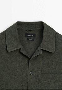 Donkergroene katoenen blouse met een klassieke kraag, een borstzak en een zwarte knoop. Zachte textuur, minimalistisch ontwerp.