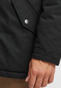 Svart isolert jakke med en jevn stofftekstur, en klaffelomme med sølvfiner, og en sømt cuff-detalj.