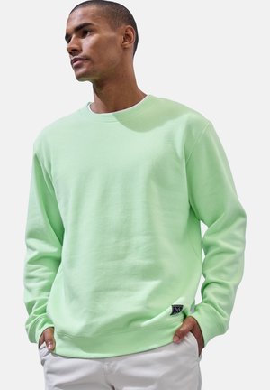 Mann trägt hellgrünes Sweatshirt mit Rundhalsausschnitt und weiße Hose, Hände in den Taschen, blickt nach links vor einfachem Hintergrund.