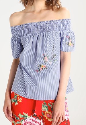 Kvinde iført en blå og hvid stribet off-shoulder top med blomsterbroderi og lyse røde bukser med blomsterprint.