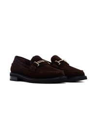 Mocasines de ante marrón con diseño de mocasín, que presentan un acento metálico en tono dorado y una suela negra plana. Textura suave y detalles de costura.