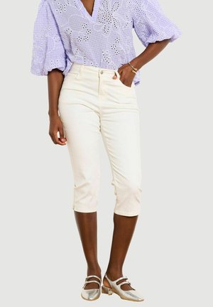 Žena nosí levanduľovú blúzku s dierkovaním a nafúknutými rukávmi, krémové nohavice po kolená, zlatý náramok a metalické Mary Jane topánky s nízkym podpätkom.