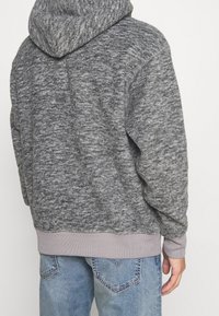 Felpa grigia in pile con texture, dotata di cappuccio, polsini e orlo a costine. Indossata sopra jeans blu, evidenziando una vestibilità comoda e un motivo discreto.