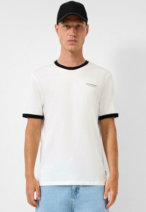 Mężczyzna w czarnej czapce, białej koszulce z czarnym obszyciem i małym napisem na piersi oraz jasnoniebieskich dżinsach, stoi na tle jednolitego tła.