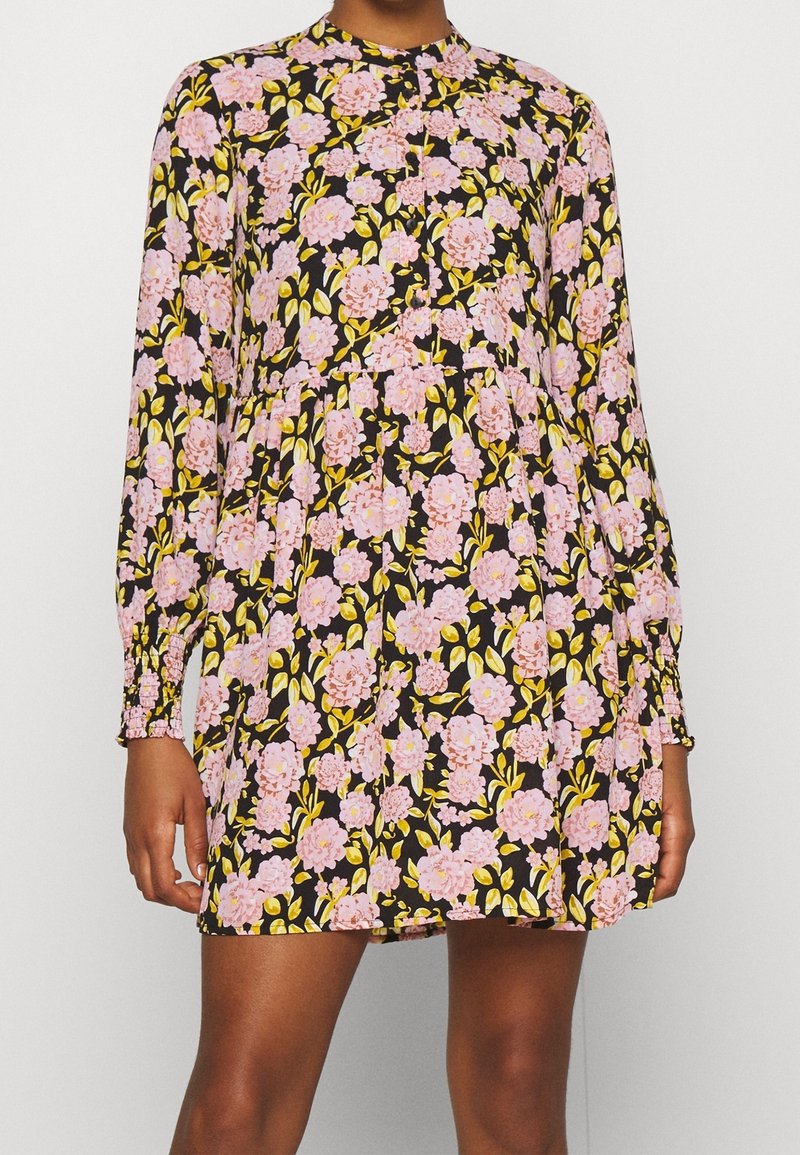 Femme portant une mini-robe noire à manches longues avec un motif floral rose et des feuilles jaune-vert.