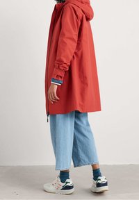 Rød vandtæt jakke med hætte, lynlåslukning og justerbare manchetter, båret over en blåstribet langærmet skjorte og lyse denimbukser.