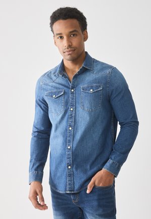 Denim košulja na dugih rukava srednje plave boje sa okovratnikom, dva džepa na grudima i srebrnim dugmadi; glatka tekstura s ležernim krojem.