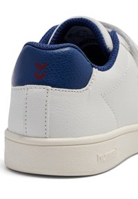 Biały skórzany sneaker z niebieskim zapiętkiem, teksturowanymi akcentami i czerwonym logo w kształcie szewrona. Posiada gumową podeszwę i wyściełany kołnierz.