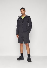 Schwarze Softshelljacke mit grauem Futter und mehreren Reißverschlusstaschen, kombiniert mit dunklen Shorts und schwarzen Sportschuhen, getragen vor einem schlichten Hintergrund.