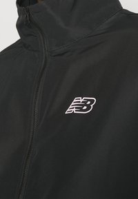 Svart vindjacka med hög krage, framzipper och en vit "NB"-logotyp på vänster bröstområde. Slät textur och lättviktsdesign.