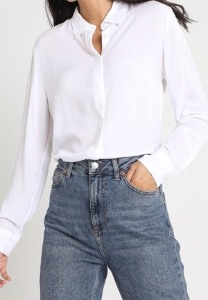 Bílá košile z hladké látky s límečkem a dlouhými rukávy, doplněná vysokým pasem modrých džínů.