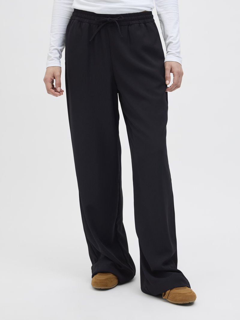 Pantalons larges noirs avec une taille élastique et un cordon de serrage réglable, fabriqués en tissu lisse, portés avec des chaussures brunes à enfiler.