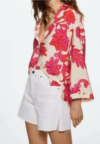 Blusa con motivo floreale, caratterizzata da fiori rosa e rossi su uno sfondo beige, abbinata a pantaloni corti in denim bianchi con orli sfrangiati e tasche.