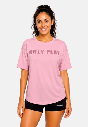 Frau mit dunklen Haaren im Pferdeschwanz, die ein pinkes "ONLY PLAY" T-Shirt und schwarze Shorts trägt, lächelt mit entspannten Händen an den Seiten.