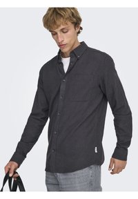 Mørkegrå skjorte med knapper foran, lavet af tekstureret stof. Har en brystlomme, blå knapper og en buet kant. Båret over en hvid skjorte.