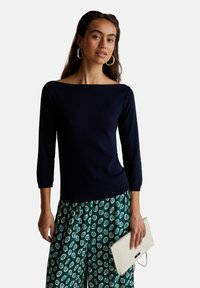 Suéter de cuello barco en azul marino con mangas tres cuartos, combinado con pantalones verdes con estampado y un bolso de mano crema. Textura suave en general.