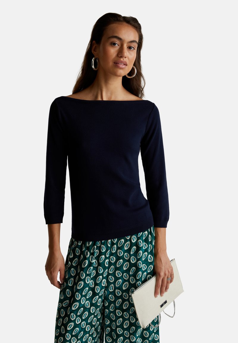 Suéter de cuello barco en azul marino con mangas tres cuartos, combinado con pantalones verdes con estampado y un bolso de mano crema. Textura suave en general.