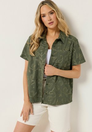 Blondynka w zielonej koszuli na krótki rękaw z motywem kwiatowym, nosząca białą bluzkę i białe szorty, pozująca na tle białej ściany.