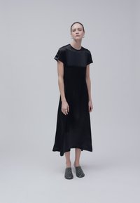 Femme debout portant une robe midi noire à manches courtes et des chaussures slip-on noires cloutées sur un fond gris uni.