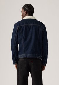 Chaqueta de mezclilla azul oscuro con un cuello de borreguito blanco, que presenta costuras visibles y detalles de botones a presión. La vista trasera muestra un diseño ajustado.