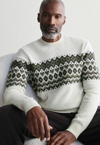 Cremefarbener Strickpullover mit einem grünen und schwarzen geometrischen Muster über der Brust und den Ärmeln, mit gerippten Bündchen und Halsausschnitt.