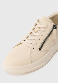 Beige leather sneaker with fabric tongue, laces, rubber sole, and a black side zipper on a plain white background.