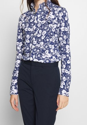 Femme portant une chemise bleue marine à fleurs boutonnée, rentrée dans un pantalon taille haute bleu marine foncé, debout devant un fond uni.