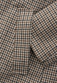 Ruskea ja valkoinen houndstooth-kangas, jolla on pehmeä rakenne, mukaan lukien lähikuva ommelluista reunoista ja näkyvästä taskusta.