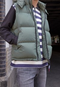 Prošívaná vesta olivově zelené barvy se zipovým zapínáním a kapsami po stranách; nošena přes námořnicky modro-bílý pruhovaný svetr a šedé džíny.